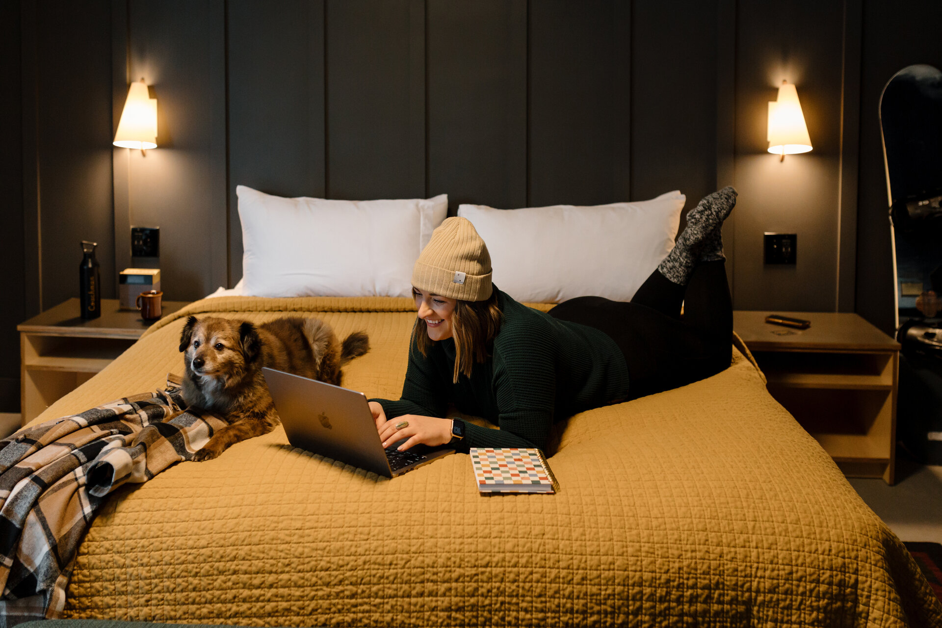 A woman and her dog on a bed with a laptop and notebook