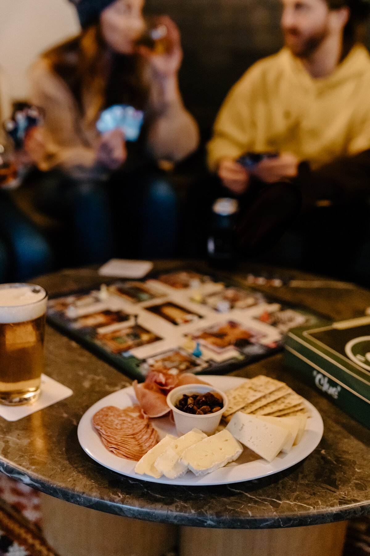 A plate of cheese, crackers, and beer on a table with people in the background