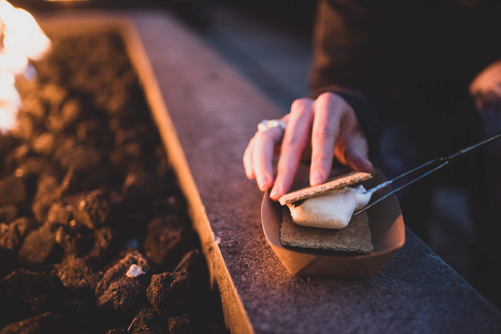 A hand holding a marshmallow on a graham cracker over a fire pit.